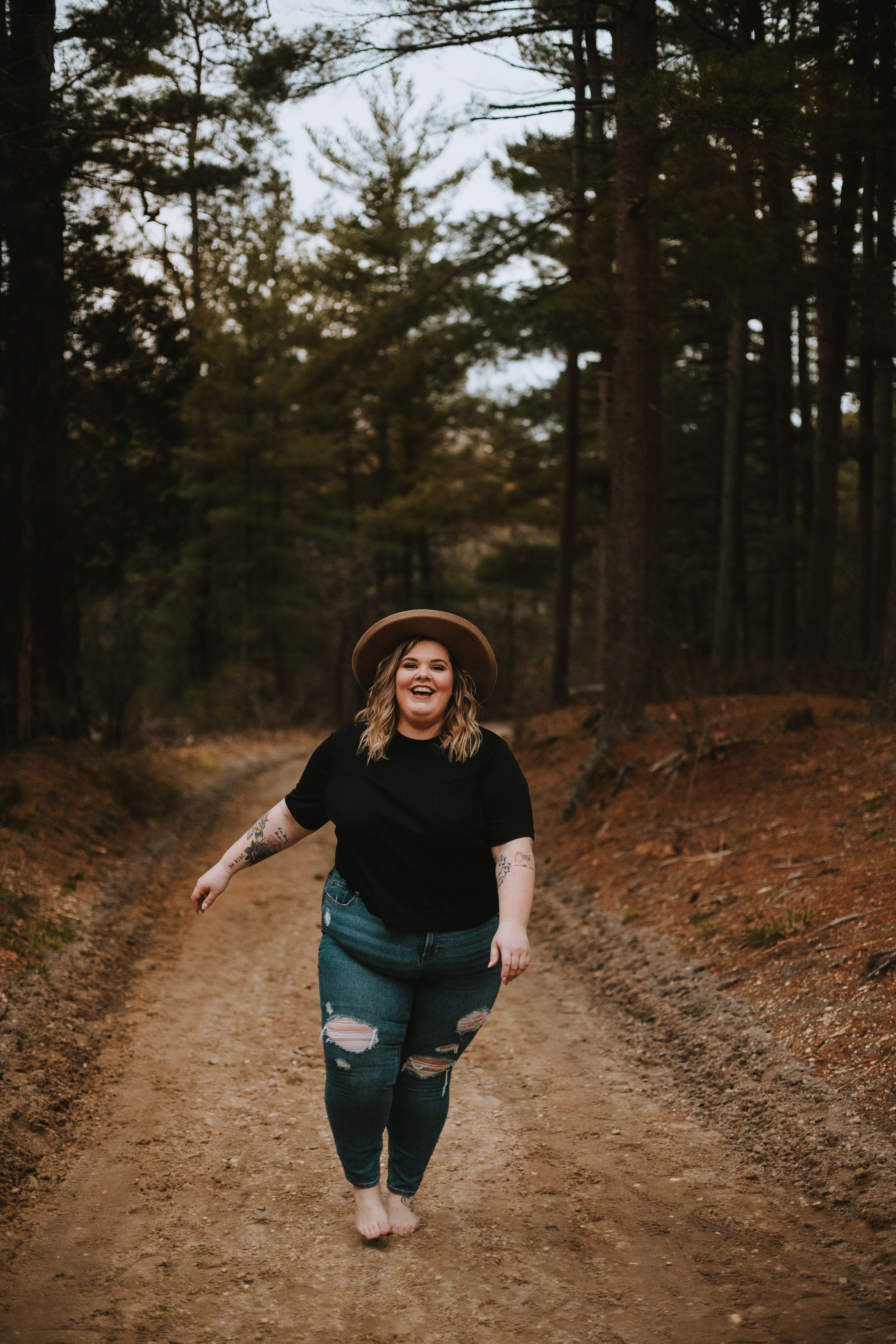 A fat blond caucasian woman is walking in a forest. She has bare feet and is wearing blue jeans, a  black t-shirt and a brown hat. She has a big smile on her face.